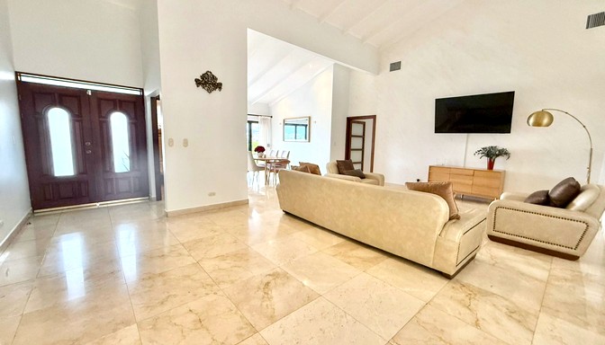 Sala de estar amplia y luminosa con pisos de mármol, muebles beige, televisor de pantalla plana en la pared y una lámpara de pie dorada; se observa la entrada principal con puerta de madera.