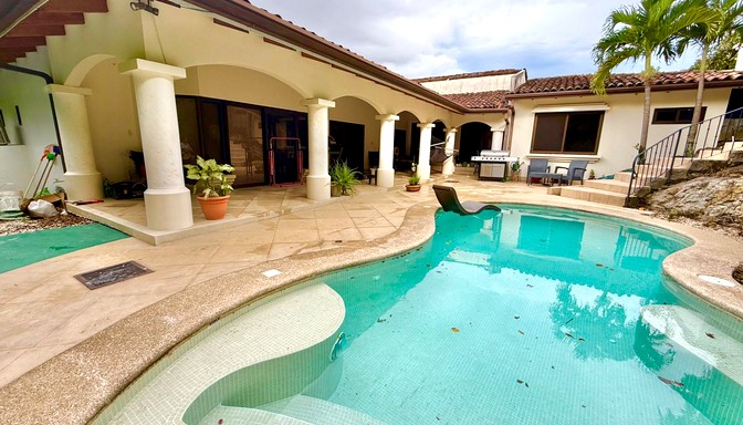Patio trasero con piscina de forma orgánica, terraza techada con columnas, plantas en macetas y área de descanso con palmeras y muebles de exterior.
