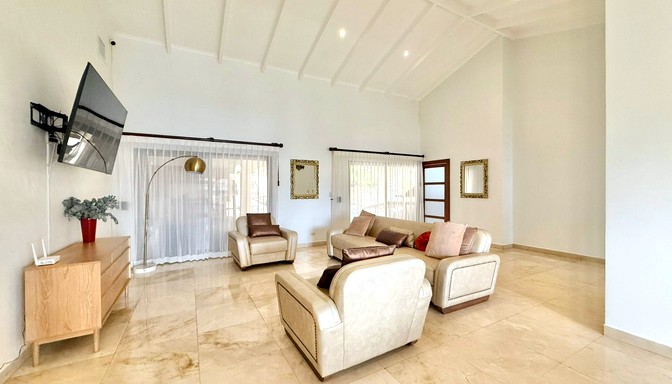 Sala de estar amplia con techo alto, sofás claros, cojines decorativos, televisor en la pared, lámpara de pie dorada y cortinas blancas translúcidas.