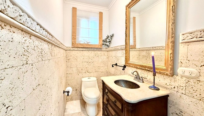 Baño pequeño con paredes de piedra clara, inodoro, mueble de madera con lavabo integrado, espejo grande con marco dorado y una vela encendida junto al grifo.