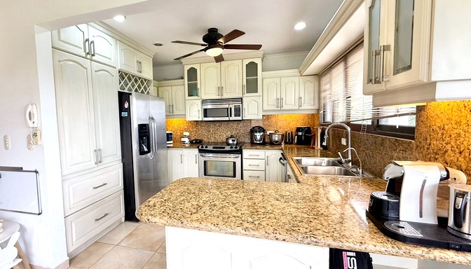 Cocina moderna de color blanco con encimeras de granito, electrodomésticos de acero inoxidable, ventilador de techo y ventanas grandes con persianas que dejan entrar luz natural.