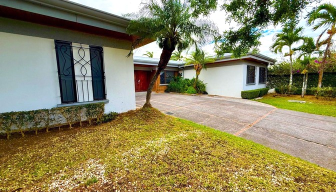 foto de casa en los laureles escazu Casa de una planta con entrada amplia, puertas de garaje blancas, jardín delantero con césped y árboles, y reja decorativa en la ventana a la derecha.